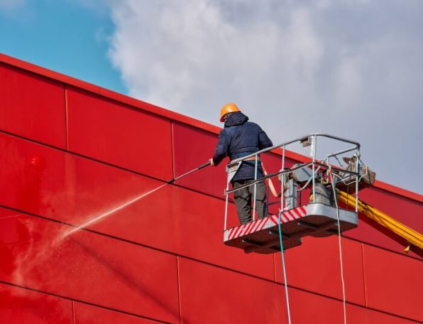 A worker in a lift cleaning a red wall with a pressure washer against a cloudy sky.